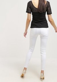 Zwarte kanttop met korte mouwen en gestructureerde stof, gecombineerd met witte skinny jeans. Model draagt zilveren hoge hakken.