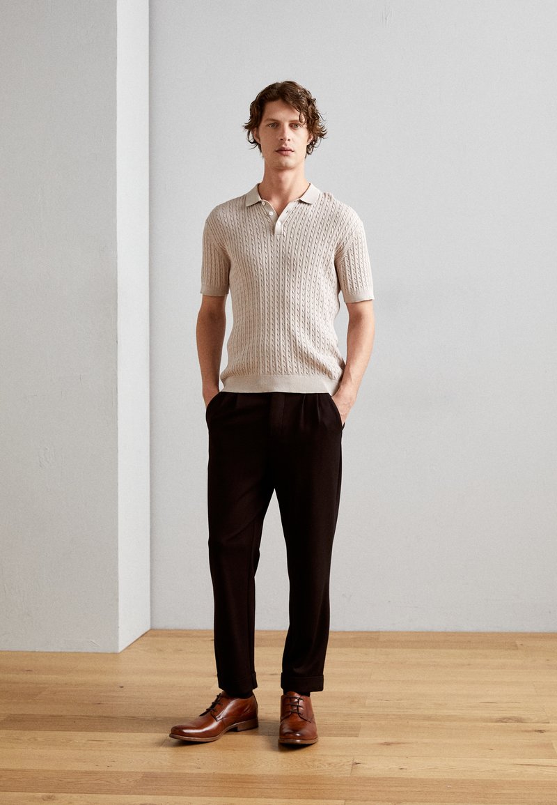 Beige Kurzarm-Polo aus Strick mit strukturiertem Muster, kombiniert mit schwarzen Hosen und braunen Lederschuhen, stehend auf einem Holzboden.