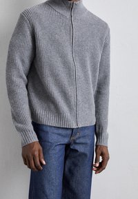 Grå ribbad zip-up tröja med hög krage, med texturerad stickad tyg och figursydda ärmslut, i kombination med blå denimbyxor.