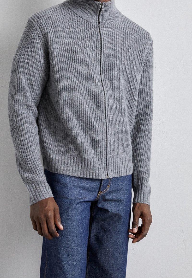 Grå ribbad zip-up tröja med hög krage, med texturerad stickad tyg och figursydda ärmslut, i kombination med blå denimbyxor.