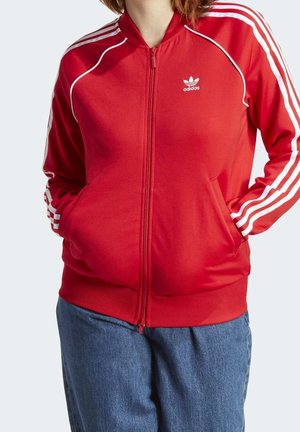 Person, die eine rote Adidas-Zipjacke mit weißen Streifen an den Ärmeln und blaue Jeans trägt, Hände in den Taschen, dargestellt vom Hals bis zu den Hüften.
