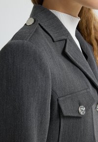 Blazer gris ajustado con hombros estructurados, cuello con muesca, botones plateados y dos bolsillos en el pecho con solapas decorativas.