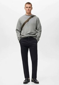 Sudadera gris de manga larga, pantalones negros y zapatos negros. El modelo está de pie con una banda de bolso marrón cruzando el cuerpo.