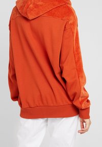 Oranje hoodie met een gestructureerd corduroy paneel op de schouders en een gladde stoffen body. Geribbelde manchetten en zoom, ontspannen pasvorm. Witte broek zichtbaar.