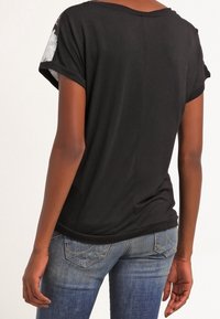 Persona vistiendo una camiseta negra de manga corta con detalles florales en las mangas y jeans azules desgastados, mostrada desde atrás.