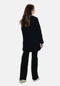 Cappotto nero oversize con maniche lunghe e linea dritta, abbinato a pantaloni neri a zampa e sneakers bianche. Texture del tessuto liscia.