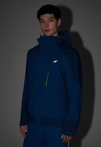 Jeune homme portant une veste à capuche bleu foncé avec un logo blanc sur la poitrine, debout devant un fond gris uni.