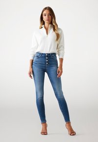 Bela rebrasta pletena jopica z zadrgo v kombinaciji z modrimi skinny jeans hlačami z visokim pasom, ki imajo pet gumbov in rahlo obrabo. Prozorni čevlji s peto.