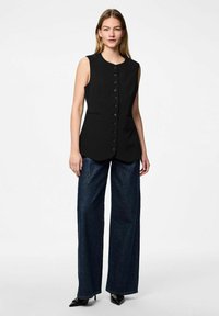 Gilet sans manches noir avec un col rond, des boutons à l'avant et des poches latérales, associé à un jean large foncé et des escarpins noirs à bouts pointus.
