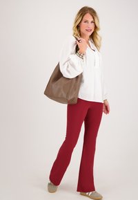 Vrouw in een witte blouse, rode broek, grijze sneakers, met een grote bruine schoudertas, glimlachend tegen een effen achtergrond.