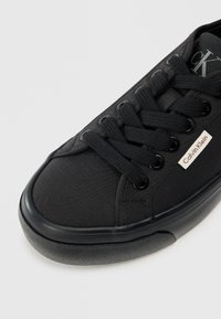 Tenisici negri din pânză cu vârf rotund, șireturi plate și talpă din cauciuc. Include eticheta cu logo-ul Calvin Klein pe lateral și material texturat.