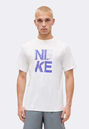 Joven con cabello rubio corto que lleva una camiseta blanca de Nike con un gran logo morado y pantalones cortos grises, de pie frente a un fondo liso.