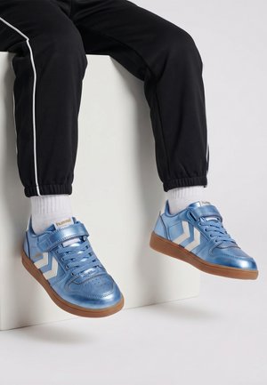 Person trägt metallisch blaue Sneaker mit weißen Chevrons, weiße Socken und schwarze Hose mit weißen Seitenstreifen, sitzt auf einem weißen Block.