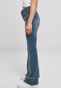 Blue flared jeans with a mid-rise waist, featuring a faded texture, five-pocket design, and contrasting stitching. Paired with white sneakers.