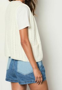 Kvinna som bär en vit kortärmad skjorta, en ärmlös krämfärgad stickad väst med fickor, och tvåfärgade blå denimshorts, sedd från sidan och bakifrån.