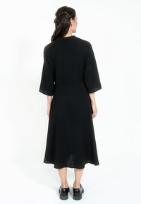 Fekete, közepes hosszúságú ruha, szűkített derékkal, bővülő szoknyával és 3/4-es ujjakkal. Simasága mellett enyhe fényességgel rendelkezik. Fekete lapos cipők.