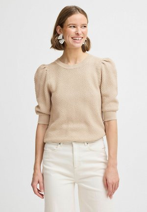 Maglione beige lavorato a maglia con motivo testurizzato, maniche a sbuffo corte e scollatura rotonda. Abbinato a pantaloni chiari a gamba larga.