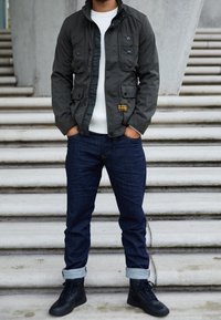 Zielona kurtka wojskowa z wieloma przednimi kieszeniami, guzikowymi detalami i wysokim kołnierzem, w połączeniu z białym swetrem z prążkowanej dzianiny i ciemnymi dżinsami.