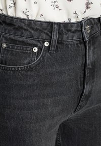 Primo piano di jeans neri in denim con tasca anteriore visibile, passante per cintura, rivetti metallici e bottone, indossati con una blusa bianca floreale infilata dentro.