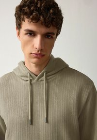 Sudadera con capucha de color caqui claro y tejido acanalado, que cuenta con cordones ajustables que tienen puntas metálicas y una superficie de tela texturizada.