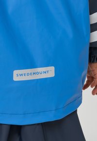 Veste imperméable bleue avec le logo "SWEDEMOUNT" en bas du dos, portée par une personne avec une main partiellement visible et une manche noire.