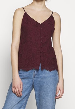 Vrouw draagt een mouwloze bordeauxrode kanten top met knopen, dunne bandjes en lichtblauwe high waist jeans.