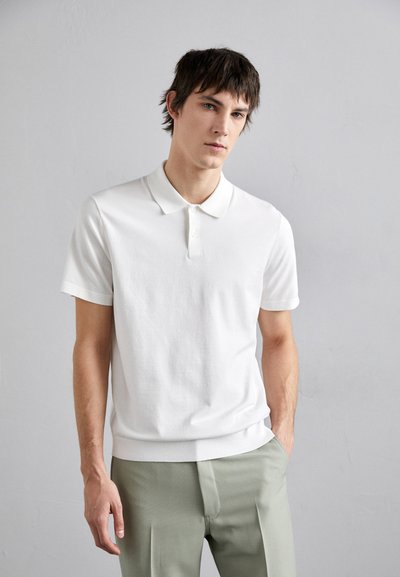 Polo shirt en coton blanc à manches courtes et patte de boutonnage à trois boutons, associé à un pantalon plissé vert clair. Texture lisse, coupe décontractée.