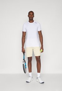 Weißes Sport-T-Shirt, beige Shorts, hält einen Tennisschläger und einen Ball, trägt Sportschuhe und weiße Socken. Einfaches Design, minimale Details.