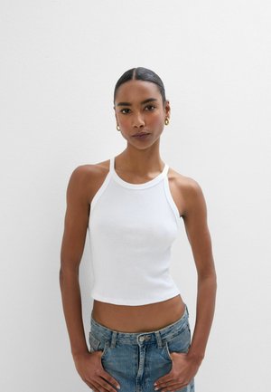 Femme aux cheveux tirés en arrière, portant des créoles dorées, un crop top blanc sans manches en maille côtelée et un jean en denim bleu, debout contre un mur blanc.