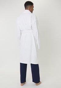 Homme debout pieds nus, portant un peignoir blanc noué à la taille sur un pantalon bleu foncé, faisant face à un fond clair et unicolore.