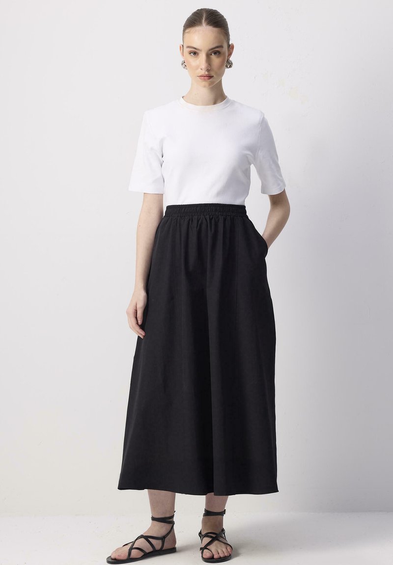 T-shirt blanc à manches courtes associé à une jupe maxi noire taille haute avec une ceinture élastique et des poches latérales. Sandales plates noires.