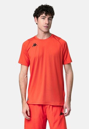 Giovane uomo che indossa una maglietta sportiva arancione brillante e pantaloncini con loghi neri Kappa, in piedi davanti a uno sfondo chiaro e uniforme.