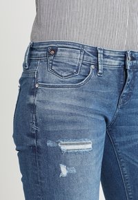 Jeans ajustados de mezclilla azul con detalles desgastados, incluyendo un pequeño desgarro y un parche de rayas verticales. Estilo clásico de cinco bolsillos.