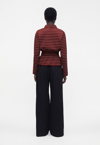Personne debout de dos, portant un haut à manches longues rayé rouge et blanc avec une taille froncée et un pantalon noir à jambes larges.