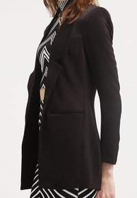 Blazer noir à la coupe ajustée, avec un col à revers et une poche avant. Tissu lisse avec une finition mate, associé à une robe géométrique en dessous.