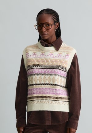 Frau mit geflochtenem Haar trägt große Brille, eine cremefarbene Strickweste mit rosa und beigen Mustern über einem braunen Cordhemd und brauner Hose.