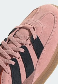 Light pink suede sneaker featuring black suede stripes, flat laces, and a textured gum rubber sole. Rear has a subtle logo detail.