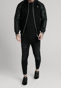 Czarna kurtka bomber z zamszowym korpusem i skórzanymi rękawami, zestawiona z czarnymi, podartymi obcisłymi dżinsami oraz czarnymi trampkami. Widoczny prosty detal z logo.