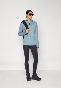 Jacket azul claro com fecho éclair, leggings pretas com painéis e sapatos de caminhada pretos. A modelo usa óculos de sol e leva uma mochila preta.