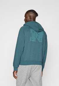 Mėlynas džemperis su apvaliu gobtuvu ir kengūros kišene, su dideliu pilku Nike logotipu nugaros pusėje, pagamintas iš minkštos, tekstūrinės medžiagos.
