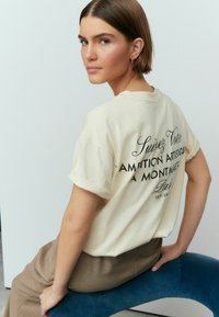 Tricou bej deschis cu mâneci răsucite, având text imprimat negru pe spate. Purtați cu pantaloni bej cu talie înaltă, așezat pe un scaun albastru.