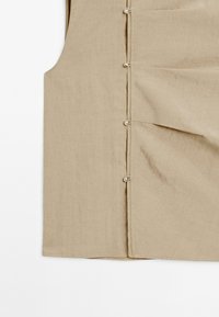 Beige Leinenweste mit leicht strukturiertem Oberflächen, ausgestattet mit Metallknöpfen an der Seite und zwei genähten Taschen.