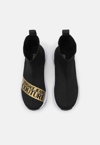 Černé slip-on tenisky s žebrovaným pleteným svrškem, bílou gumovou podrážkou a zlatým logem na boku. Hladký povrch, minimalistický design.