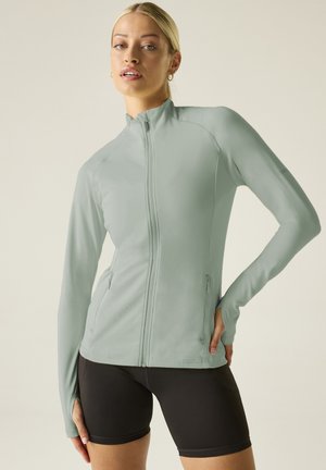 Young woman wearing a light green zip-up athletic jacket with thumb holes and black cycling shorts against a plain background.