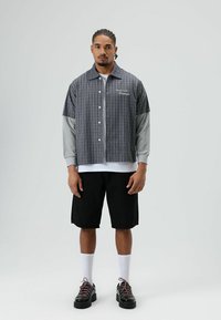 Grijs-blauw geruit overhemd met korte mouwen en knopen, gedragen over een grijs shirt met lange mouwen, gecombineerd met zwarte korte broek en zwarte schoenen.