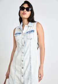 Jeans ärmlös klänning i ljusblått med knappstängning, två framfickor och krage. Modell som håller en brun handväska och bär svarta solglasögon.