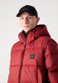 Crvena puffer jakna s visokim ovratnikom, kvadratnim dijelovima i crnom logotipskom naljepnicom. Uparen s crnom kapicom sa bijelim tekstom.