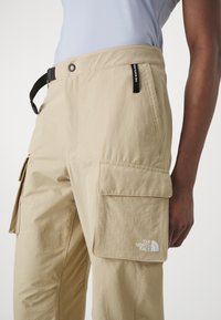 Närbild av beige cargobyxor med sidofickor, ett svart justerbart bälte och The North Face-logotypen på ficklocket.