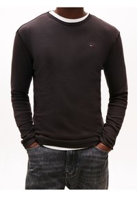 Man wearing black long-sleeve sweatshirt with small logo on chest, white undershirt showing at collar, and dark washed jeans with hands in pockets.