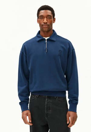 Mann trägt ein marineblaues Sweatshirt mit Viertel-Reißverschluss und kleinem gesticktem Logo auf der Brust sowie schwarze Jeans, steht vor einem weißen Hintergrund.
